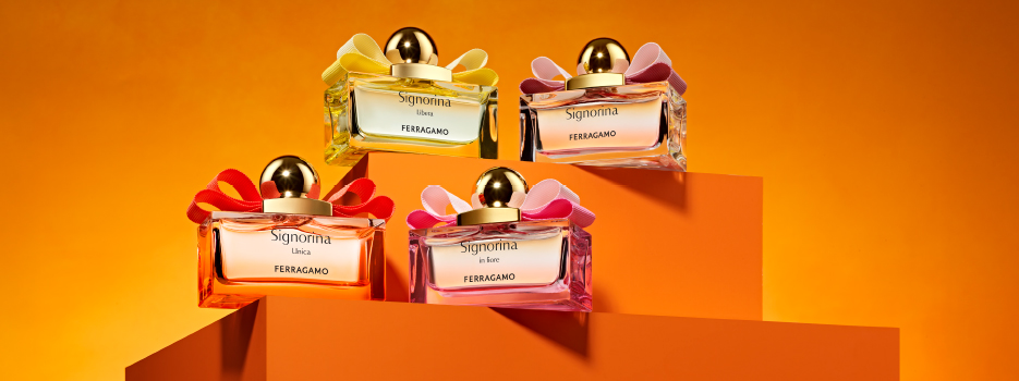 Vier Parfumflakons der 'Signorina'-Kollektion von Ferragamo stehen auf orangefarbenen Podesten vor einem warmen, orange leuchtenden Hintergrund. Jeder Flakon ist mit einer goldenen Kugelkappe und einer farbigen Schleife verziert. Die Düfte heißen 'Signorina Libera', 'Signorina', 'Signorina in Fiore' und 'Signorina Unica'. Die Szene wirkt elegant, feminin und farbenfroh.
