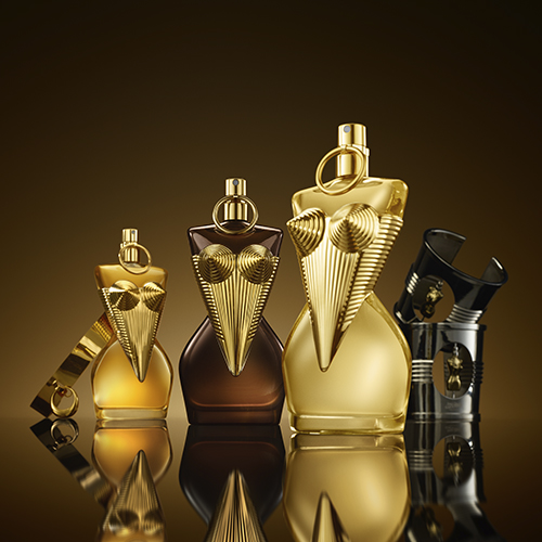 Vier goldene und bronzefarbene Parfumflakons mit kunstvollem, maskenähnlichem Design und spitz zulaufender Form, stehend auf spiegelnder Fläche vor dunklem Hintergrund.