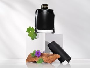 Montblanc Legend Eau de Parfum in einem glänzend schwarzen Flakon mit goldener Beschriftung. Präsentiert auf einem weißen Podest, umgeben von Holzstücken, einem violetten Blümchen und grünen Blättern – Hinweis auf die natürlichen Duftbestandteile.