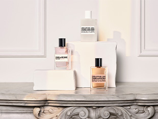 Drei Parfumflakons von Zadig & Voltaire stehen auf weißen Podesten vor einer eleganten, klassischen Wandverzierung. Die Flakons tragen schwarze Schriftzüge, einer ist weiß, die anderen zwei transparent mit rosé- und bernsteinfarbenem Inhalt. Sie sind auf einer Marmoroberfläche arrangiert und in sanftem Licht inszeniert.