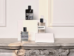 Drei Parfumflakons von Zadig & Voltaire für Herren stehen auf weißen Podesten vor einer hellen, klassisch verzierten Wand. Die Flakons sind in Schwarz, Grau und Transparent mit dunkler Flüssigkeit gehalten und tragen schwarze Schriftzüge. Sie sind auf einer eleganten Marmorfläche arrangiert und in weiches Licht getaucht.